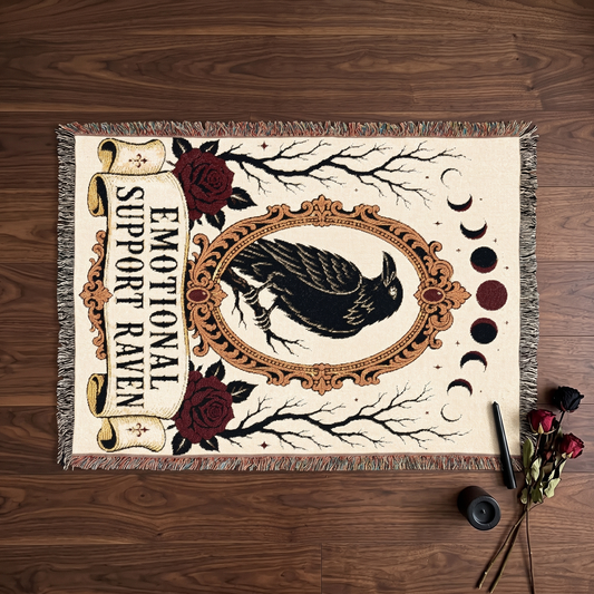 Overhead flatlay of the gothic raven tarot woven blanket on dark walnut, showing the full design with banner and multicoloured fringe