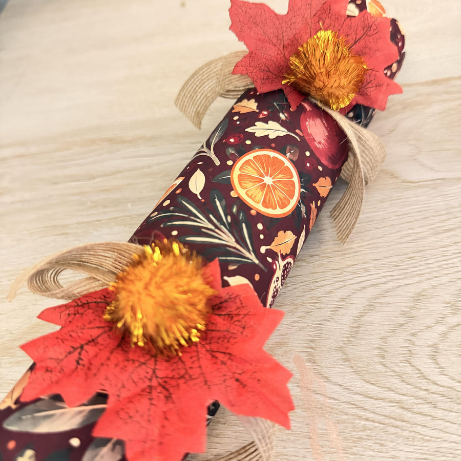 Close-up of a red maple leaf, gold tinsel pom pom and burlap bow on a burgundy citrus print cracker