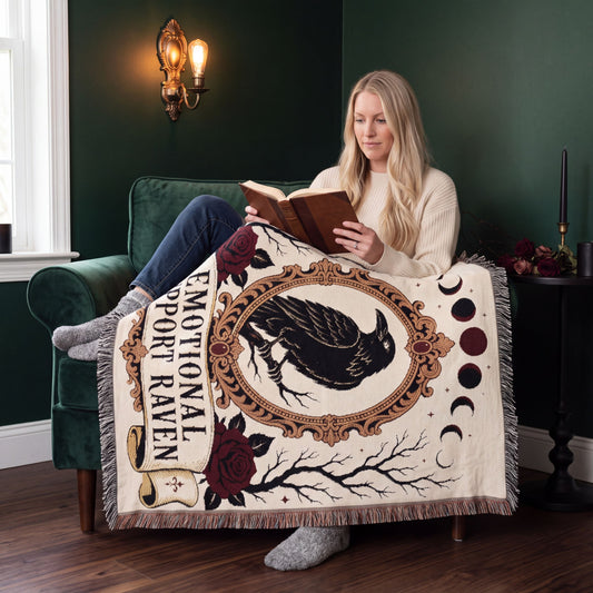 Woman reading in a green velvet armchair under the Emotional Support Raven woven blanket, gothic tarot style cotton throw