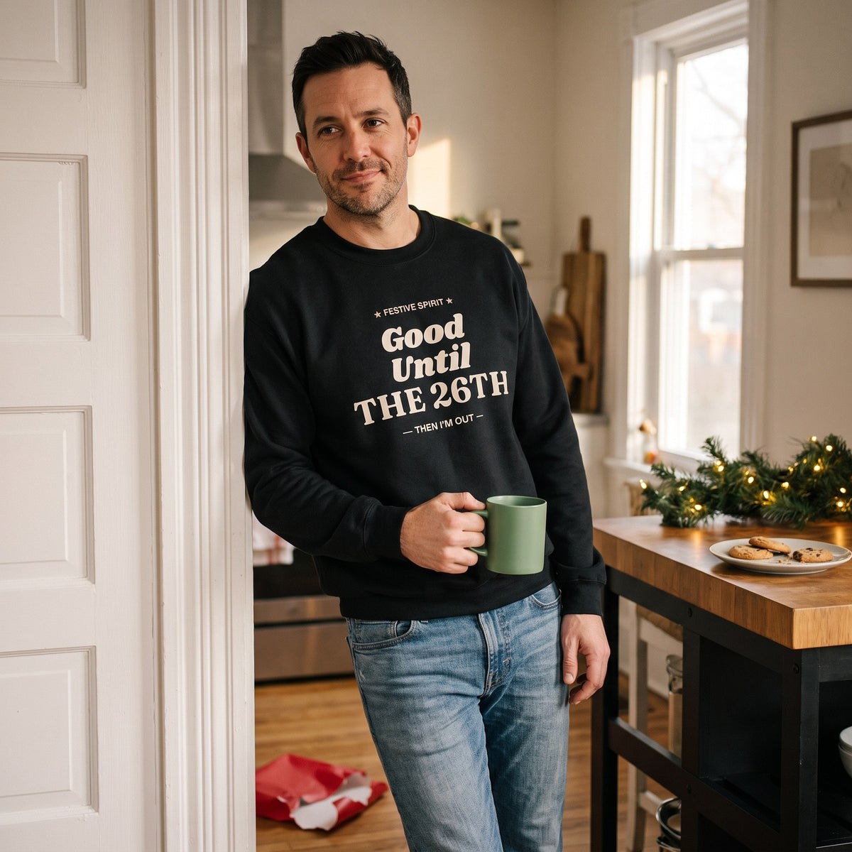 Man leaning in a kitchen doorway with a mug in the Black Good Until the 26th sweatshirt