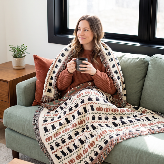 Woman wrapped in the Haunted Heirloom Fair Isle woven blanket, cream ground with charcoal ghosts, black cats and rust pumpkins, standing by a window in a mid tone living room