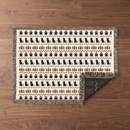 Overhead flatlay of the Haunted Heirloom woven blanket laid open on a walnut floor, showing the full Fair Isle pattern bands of ghosts, cats and pumpkins with multicoloured fringe