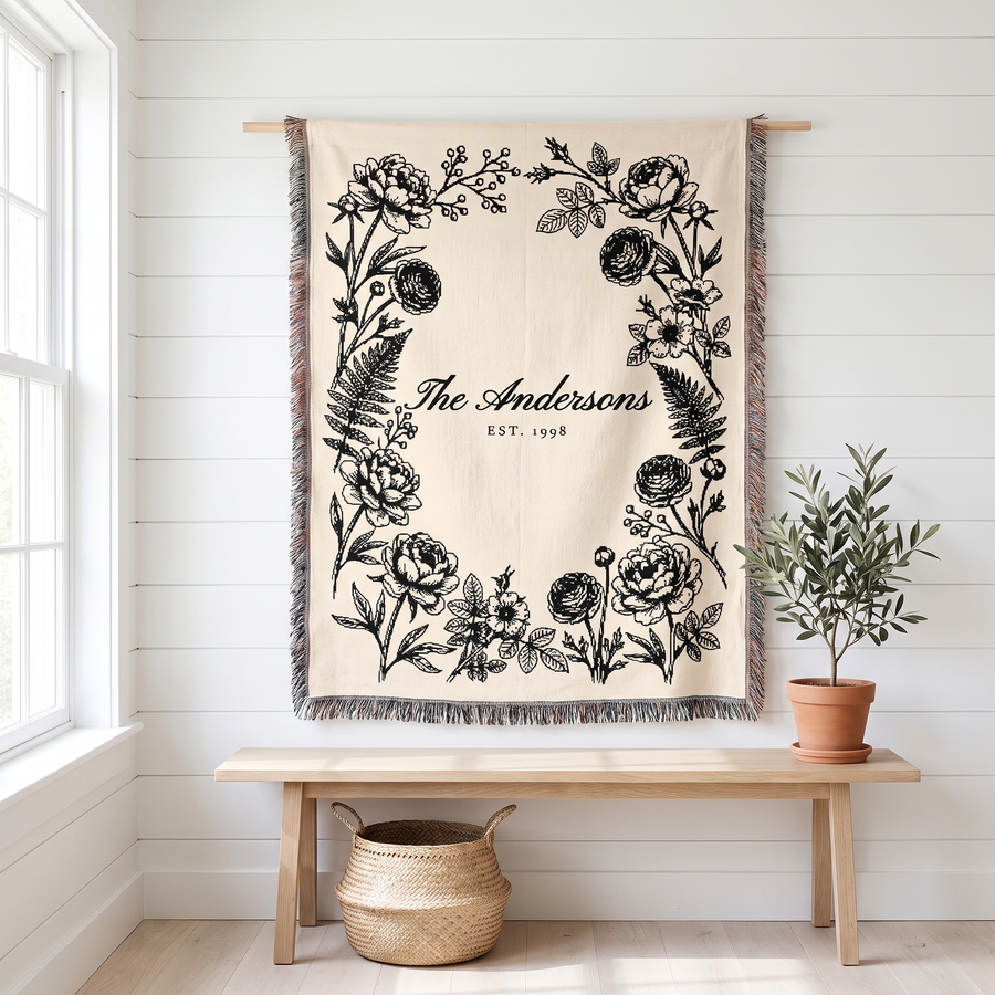 Personalized family name woven blanket, black botanical on cream cotton, hanging on a white wall