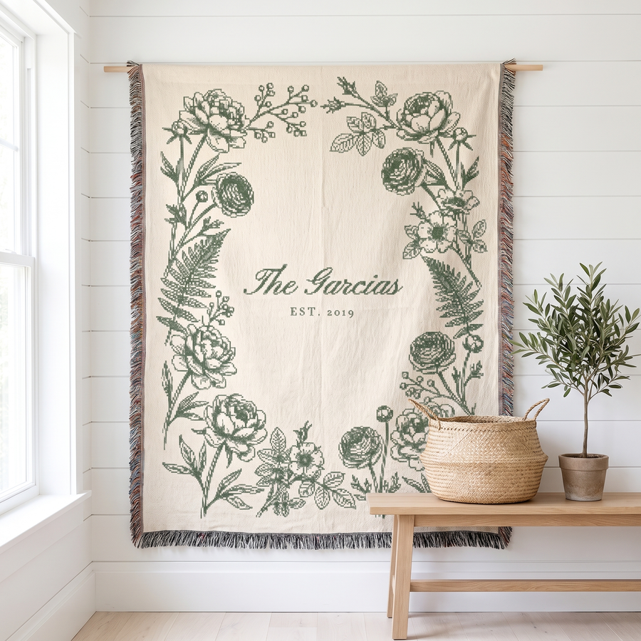Personalized family name woven blanket, sage botanical on cream cotton, hanging on a white wall