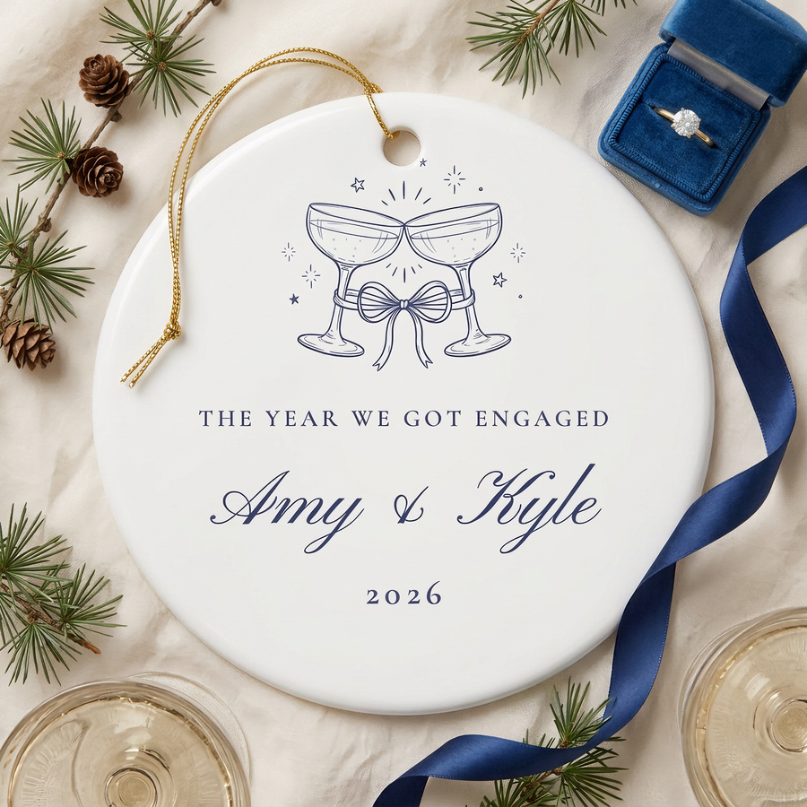 Champagne toast engagement ornament on linen with pine, champagne coupes and an engagement ring box