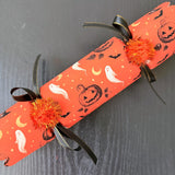Six Halloween party crackers lined up side by side with scalloped ends and orange pom poms on a dark table