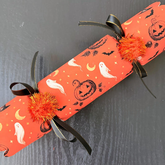 Six Halloween party crackers lined up side by side with scalloped ends and orange pom poms on a dark table