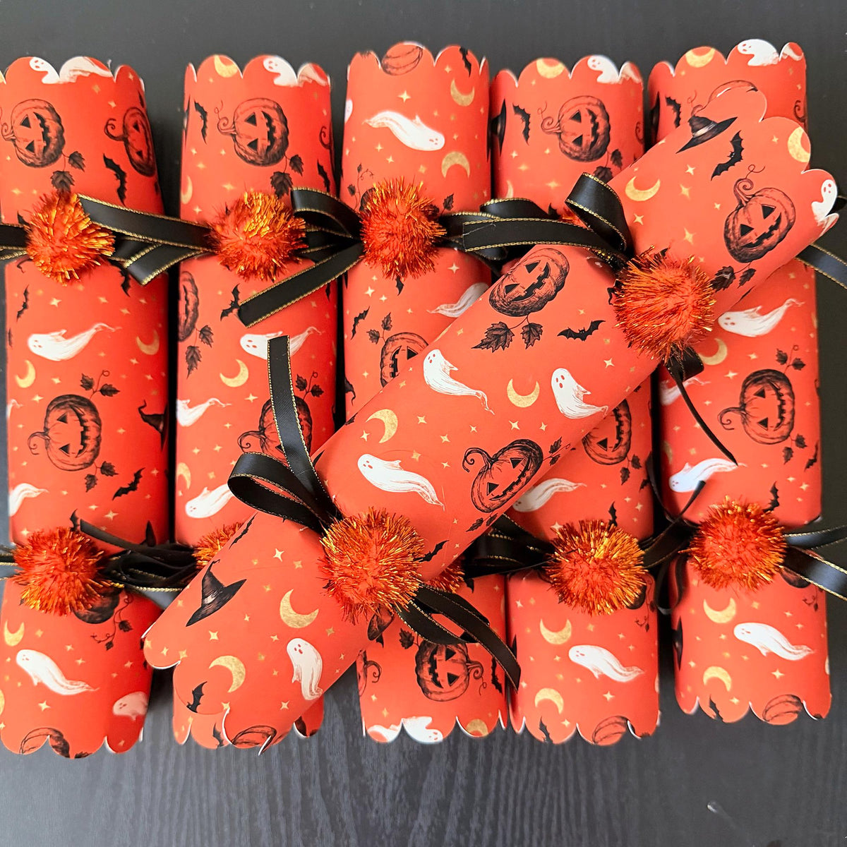 Single Halloween cracker with snap on a dark wood table, tied with black and gold-edged ribbon