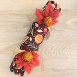 Single Mulled and Merry cracker with snap on a pale wood table, tied with burlap ribbon