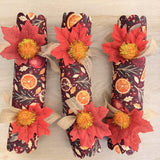 Three handmade holiday crackers side by side with scalloped ends, red maple leaves and gold tinsel pom poms