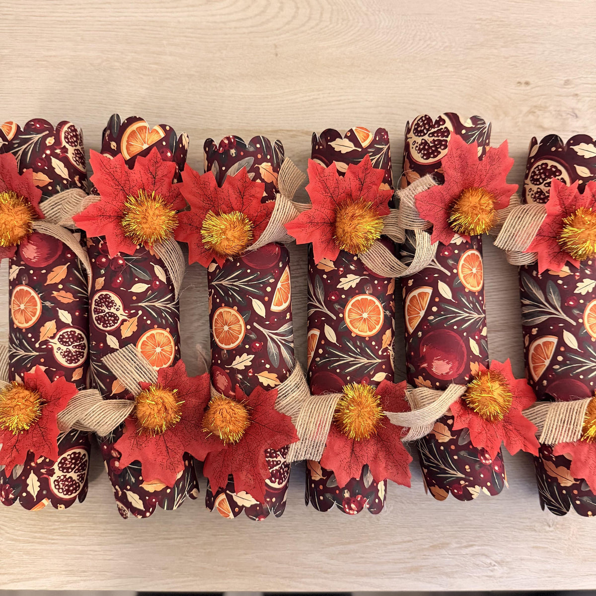 Top-down view of six burgundy crackers with orange slice and pomegranate print, burlap ribbon and red leaves