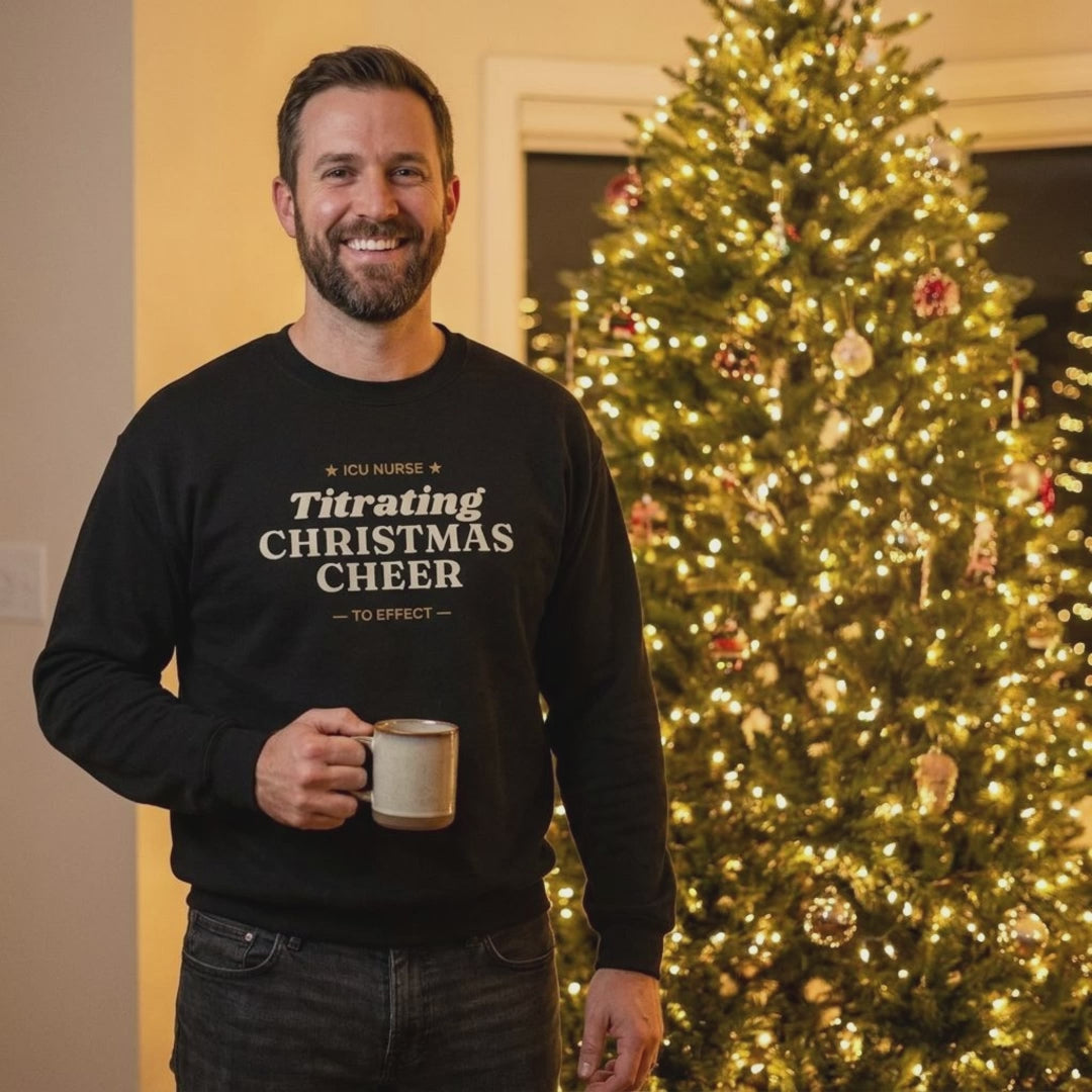 Christmas tree lights twinkling behind a man in the Black ICU nurse Titrating Christmas Cheer sweatshirt
