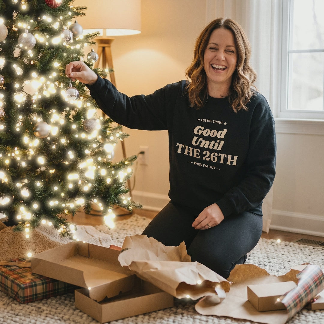 Woman trimming the Christmas tree in the Black Good Until the 26th sweatshirt with twinkling lights